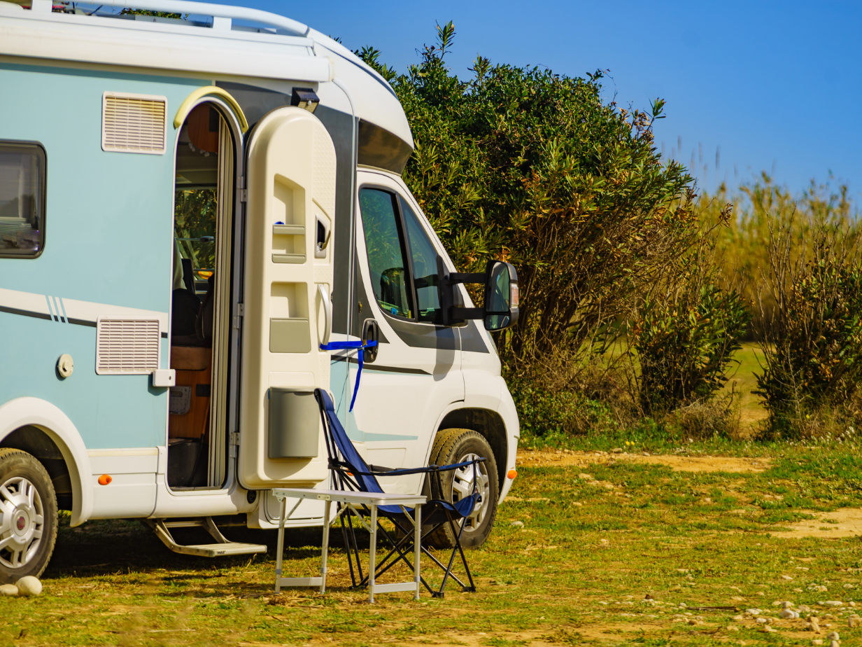 Camper met open deur op een zonnige kampeerplaats, met twee kampeerstoelen naast het voertuig