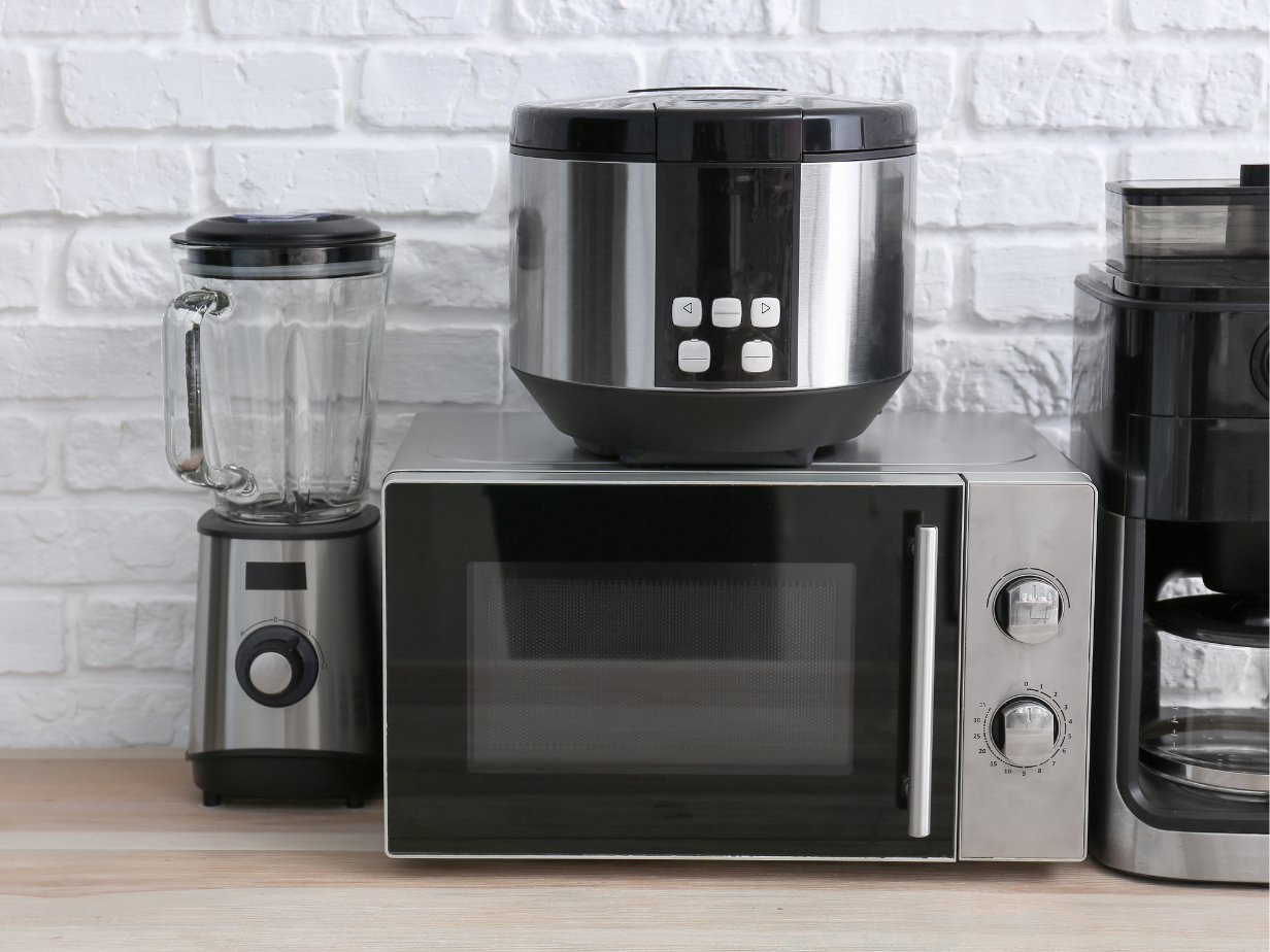 Verschillende elektrische keukenapparaten, waaronder een magnetron, blender, rijstkoker en koffiezetapparaat, op een keukenwerkblad