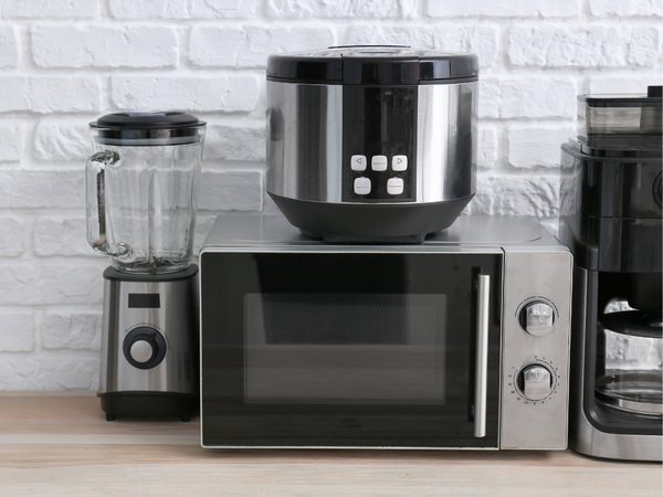 Verschillende elektrische keukenapparaten, waaronder een magnetron, blender, rijstkoker en koffiezetapparaat, op een keukenwerkblad