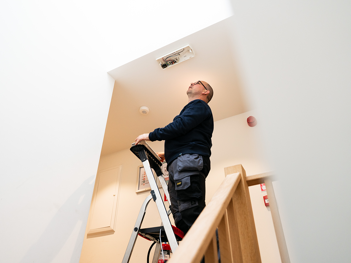 Technieker voert een keuring uit van de noodverlichting in een traphal met behulp van een ladder.