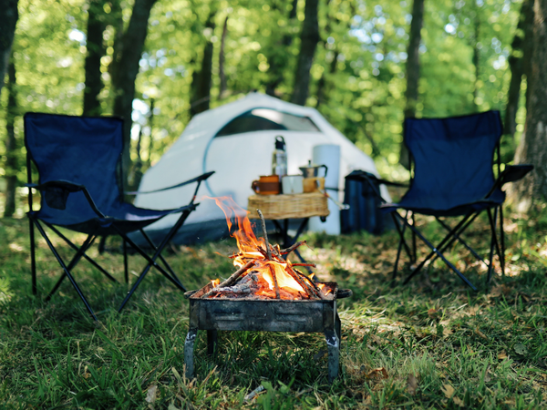 Brandend kampvuur in een vuurkorf tussen twee kampeerstoelen, met een tent op de achtergrond in een bosrijke omgeving